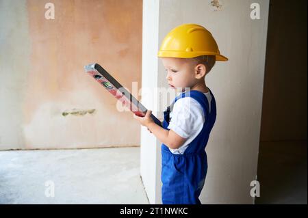 Superficie della parete di controllo per bambini con livella a bolla d'aria nell'appartamento. Bambino in tute da lavoro e casco di sicurezza da costruzione utilizzando strumenti di livello durante il lavoro di ristrutturazione a casa. Foto Stock