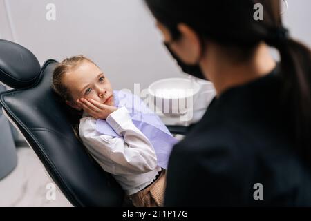 Ritratto di una bambina dall'aspetto preoccupata sdraiata sulla sedia dentale davanti al medico che si lamenta di mal di denti e che tiene la mano sulla guancia, guardando una donna Foto Stock