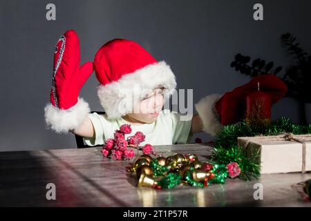 Un bambino piccolo con cappello di Babbo Natale e guanti rossi con regali di Natale è seduto al tavolo di Capodanno. Festeggiamo il Natale. Foto Stock