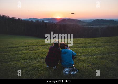Coppia seduta in cima a una collina che si gode una serata romantica. Guy vola un drone e registra un tramonto rosso mozzafiato. Foto Stock