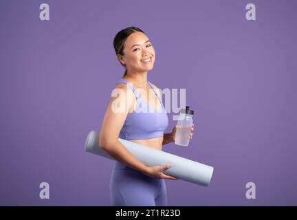 Felice donna asiatica con tappetino yoga e bottiglia d'acqua, in posa su sfondo viola Foto Stock