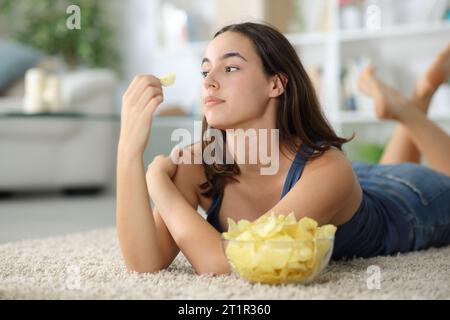 Una donna pensiva che guarda lontano a mangiare patatine fritte sdraiata per terra a casa Foto Stock