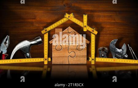 Concetto di miglioramento della casa. Casa in legno composta da pezzi di puzzle e righello pieghevole, su un banco da lavoro in legno con molti attrezzi da lavoro. Foto Stock