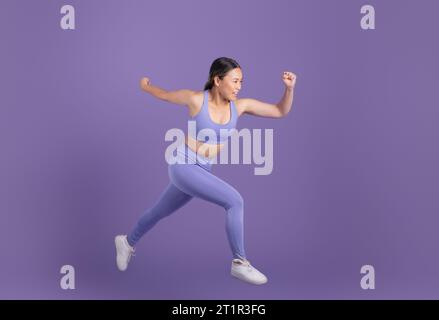 Giovane donna asiatica in abbigliamento sportivo che corre su sfondo viola studio, spazio libero Foto Stock