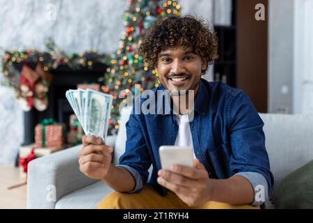Ritratto di un uomo gioioso a Natale a casa, ispanico che guarda la macchina fotografica seduto sul divano vicino all'albero delle vacanze, con denaro contante dollari e telefono, il vincitore ha vinto un vero premio utilizzando l'app sullo smartphone. Foto Stock