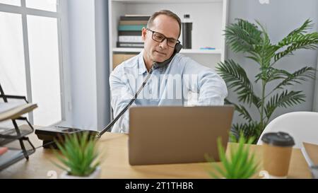 Uomo d'affari di mezza età rilassato che si occupa di una conversazione telefonica seria mentre lavora al suo notebook in ufficio Foto Stock