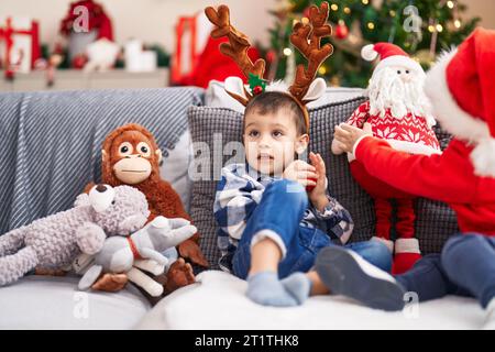 Adorabile bambino che indossa orecchie di renna seduto sul divano vicino all'albero di natale a casa Foto Stock