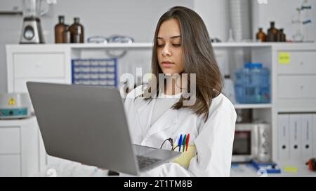 Giovane donna ispanica focalizzata, una bella scienziata al lavoro, che utilizza laptop e microscopio per la ricerca medica in laboratorio, mostrando un rilassato, ma degrado Foto Stock