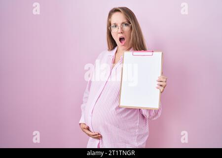 Giovane donna incinta che tiene un clipboard in faccia shock, sembra scettica e sarcastica, sorpresa con la bocca aperta Foto Stock