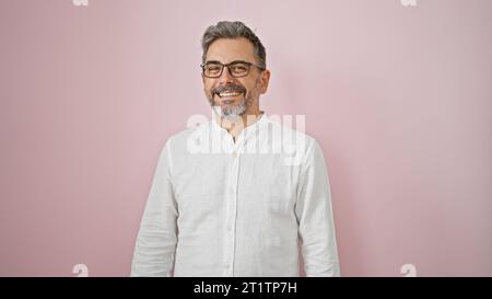 Uomo ispanico dai capelli grigi sicuro di sé che sorride con un sorriso affascinante, indossa occhiali, si erge su uno sfondo rosa isolato e irradia positività Foto Stock