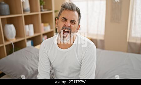 Giovane uomo ispanico esausto con i capelli grigi, che sbadiglia a bocca aperta mentre si siede su un letto accogliente, che lotta per svegliarsi in camera da letto Foto Stock