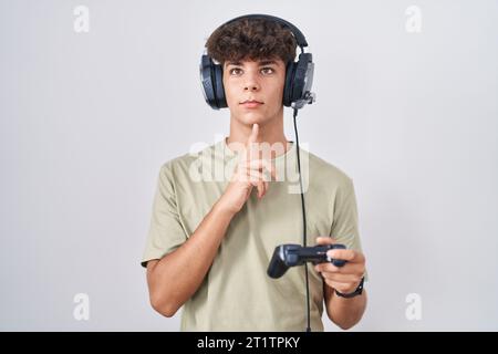 Teenager ispanico che gioca con il controller di controllo si concentra sul dubbio con il dito sul mento e lo sguardo rivolto verso l'alto a chiedersi Foto Stock