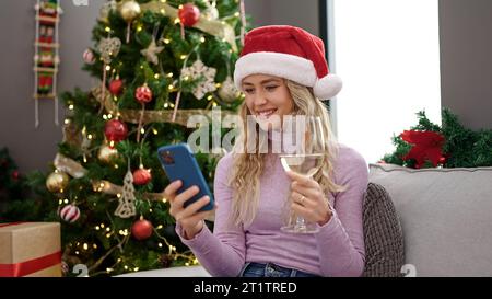 Giovane bionda che festeggia il natale con lo smartphone bevendo vino a casa Foto Stock