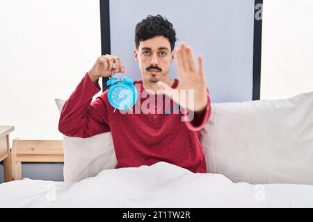 Uomo ispanico che tiene la sveglia nel letto con la mano aperta facendo segnale di stop con espressione seria e sicura, gesto di difesa Foto Stock