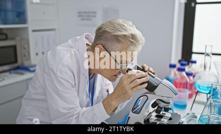 Scienziata senior con i capelli grigi dedicata immersa in un intenso lavoro di ricerca sul microscopio a livello di dottorato in laboratorio Foto Stock