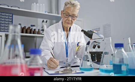 Nel cuore della scienza, una scienziata anziana dai capelli grigi, che lavora sodo nel suo laboratorio, prende appunti meticolosamente mentre pesa attraverso un micr Foto Stock