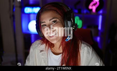 Streaming live accattivante, giovane donna che filtra rossa brillante, sorridente, che abbraccia i giochi high-tech in una stanza accogliente, con cuffie accese Foto Stock