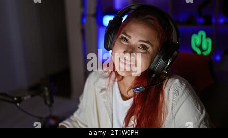 Streaming live accattivante, giovane donna che filtra rossa brillante, sorridente, che abbraccia i giochi high-tech in una stanza accogliente, con cuffie accese Foto Stock