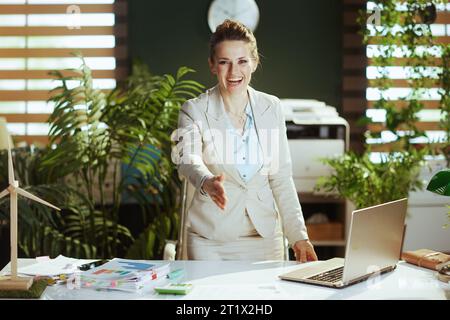 Ritratto di una donna moderna e sorridente proprietaria di una piccola impresa in un leggero abito da lavoro in un moderno ufficio verde con un laptop che dà la mano per una stretta di mano. Foto Stock