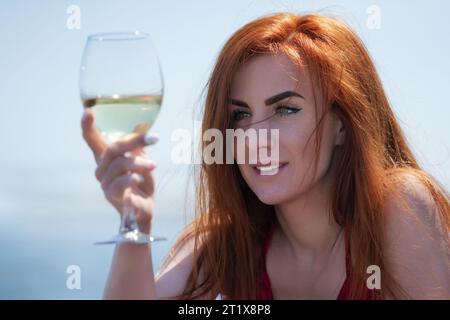 Ritratto ravvicinato Happy Redhead Woman ha alzato un bicchiere di vino bianco a livello degli occhi e lo sta guardando con attenzione. Donna sorridente in abito rosso che beve Foto Stock