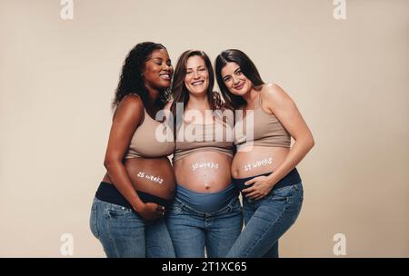 Gruppo diversificato di donne incinte nel terzo trimestre in piedi insieme in uno studio. Sorridono e mostrano i loro piccoli urti. Indossa jeans e su Foto Stock