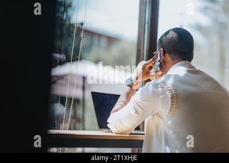 Uomo di colore, di sesso maschile, che ha telefonato mentre lavora sul computer portatile seduto alla scrivania accanto alla finestra dell'ufficio in una giornata di sole Foto Stock