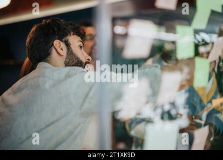 Diversi gruppi aziendali collaborano in uffici aziendali. Il team multirazziale condivide le conoscenze, scrivendo appunti adesivi su una parete di vetro. Foto Stock