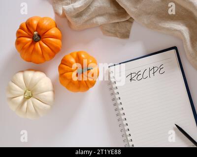 Concetto di ricette di zucca. Zucche autunnali di colore bianco e arancione su sfondo bianco con un quaderno e una penna con la parola ricetta. Vista dall'alto. Foto Stock