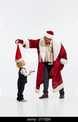 Buon Natale. Buffo padre, amico, vestito come Babbo Natale, divertiti con il piccolo bambino sudato, il sole sullo sfondo bianco dello studio. Foto Stock