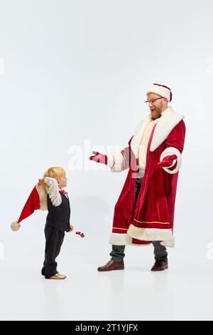 Buon Natale. Padre, uomo, vestito come Babbo Natale che si diverte con il piccolo bambino sudato, sole con cappello rosso su sfondo bianco da studio. Copia spazio. Foto Stock