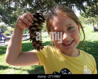 La ragazza carina e allegra che tiene in mano un cono di pino sorride. Foto Stock