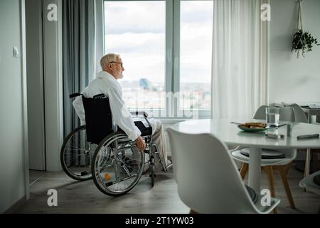 Uomo anziano in sedia a rotelle che passa del tempo da solo in appartamento. Concetto di solitudine e dipendenza dei pensionati. Foto Stock