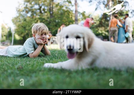 Bambini che giocano con un piccolo cucciolo a una festa in giardino per tutta la famiglia. Ritratto di un bambino sdraiato sull'erba con il cucciolo di Golden retriever. Foto Stock