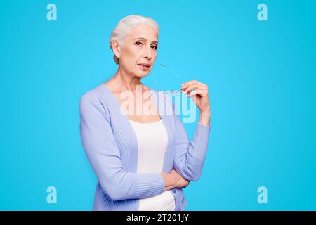 Bella bella vecchia signora dai capelli grigi che indossa casual, regge occhiali, pensa, rifletta. Isolato su sfondo viola pastello Foto Stock