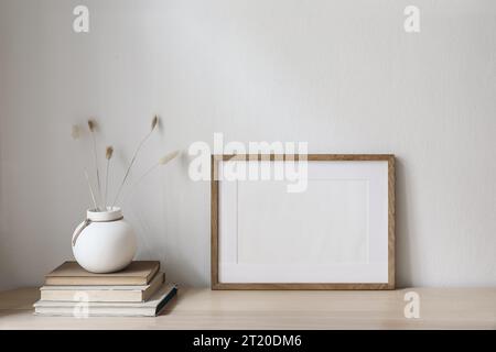 Mockup orizzontale con cornice in legno vuota sul tavolo. Elegante vaso rotondo in ceramica con coda di coniglio asciutta, erba ovatus Lagurus e vecchi libri. Muro bianco Foto Stock