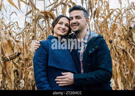 Una coppia felice innamorata si trova in autunno vicino a un campo di grano. Un uomo con una camicia e una giacca abbraccia una donna con un cappotto Foto Stock