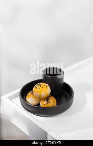 Pasticceria cinese cosparsa di semi di sesamo nero sulla parte superiore e servita con tè caldo in una tazza di ceramica nera. Foto Stock