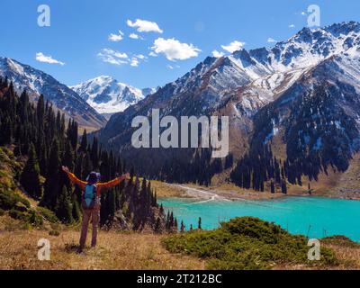 Un turista con una giacca e un cappello arancioni sta facendo un'escursione sulle montagne che si affacciano su uno splendido lago turchese. Escursioni nelle Highlands. La gioia di Foto Stock