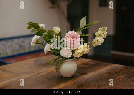 Una natura morta caratterizzata da un vaso di vetro colorato con un vivace bouquet di fiori assortiti posti su un tavolo di legno di fronte a una parete piastrellata testurizzata Foto Stock