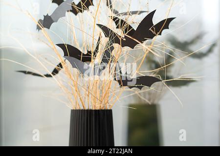 Halloween. Concetto. Vacanze autunnali. Pipistrelli spaventosi fatti di carta nera, su rami asciutti in un vaso nero. Sullo sfondo della finestra. Riflessione. Foto Stock