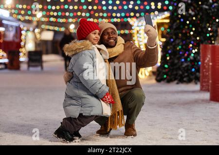 Ritratto a figura intera di Happy Black padre e figlia che scattano foto selfie all'aperto insieme al mercatino di Natale, spazio fotocopie Foto Stock