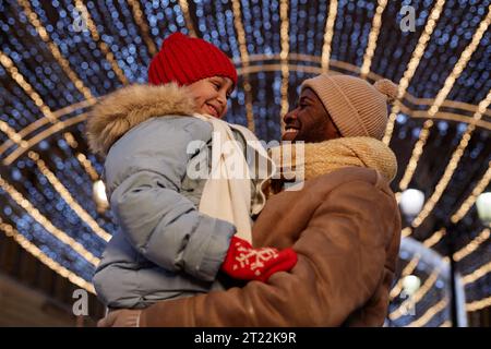 Vista laterale del padre e della figlia che si abbracciano sotto le decorazioni delle luci natalizie Foto Stock