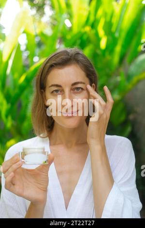 Foto verticale ravvicinata del volto di una donna di mezza età. Una donna applica la crema sotto gli occhi con movimenti massaggianti e tenendo in mano un vaso. Foto Stock