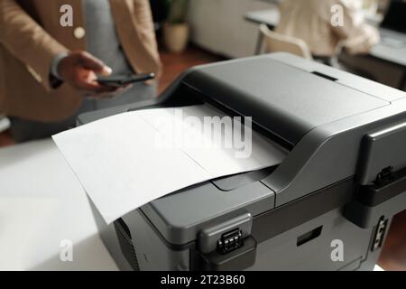 Macchina xerox in plastica grigia con documento di carta su vassoio in piedi sul luogo di lavoro del responsabile dell'ufficio o del contabile contro l'uomo con lo smartphone Foto Stock