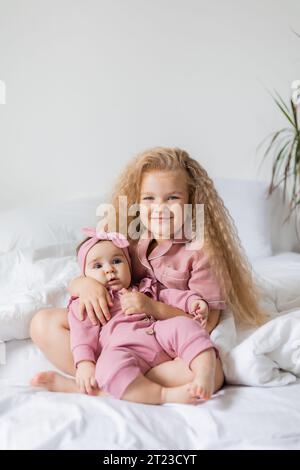 Graziosa ragazza riccia in pigiama rosa tiene un bambino tra le braccia mentre è seduto a letto Foto Stock