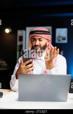 Un arabo impegnato in una videoconferenza con un collega sul suo smartphone. Utilizza sapientemente la tecnologia wireless per il lavoro e la ricerca, dimostrando le sue capacità di comunicazione digitale. Foto Stock