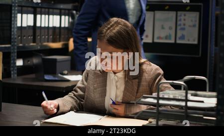 Una donna ufficiale di legge riceve informazioni cruciali sulla rete fissa e condivide nuovi indizi del caso con il detective afroamericano. Investigatore caucasico che risponde al telefono nella sala degli incidenti. Scatto con treppiede. Foto Stock