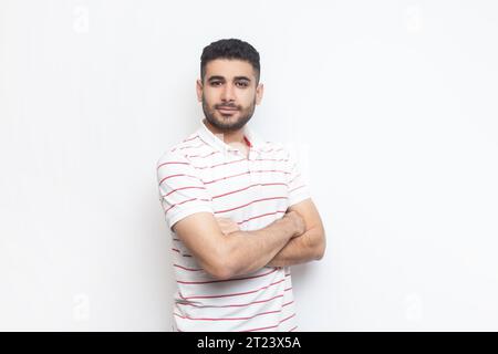 Ritratto di un uomo barbato, positivo e sicuro di sé, che indossa una t-shirt a righe con le mani incrociate e guarda la fotocamera. Riprese in studio in interni isolate su sfondo grigio. Foto Stock