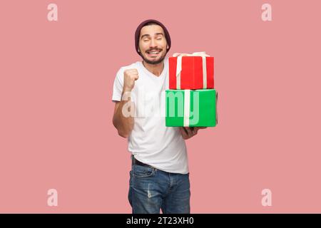 Ritratto di un uomo barbuto soddisfatto estremamente felice in T-shirt bianca e cappello di fagiolo in piedi che regge una scatola regalo, il pugno stringato, per festeggiare. Riprese in studio per interni isolate su sfondo rosa. Foto Stock