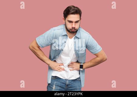 Ritratto di uomo barbuto in camicia casual in piedi sente mal di stomaco, soffre di indigestione e crampi addominali dopo aver mangiato prodotto viziato. Riprese in studio per interni isolate su sfondo rosa. Foto Stock